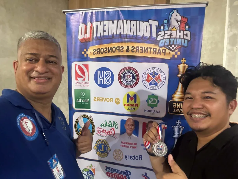 A young medalist holding up a Chess United medal beside a club organizer in front of the Tournament 1.0 partners and sponsors banner.