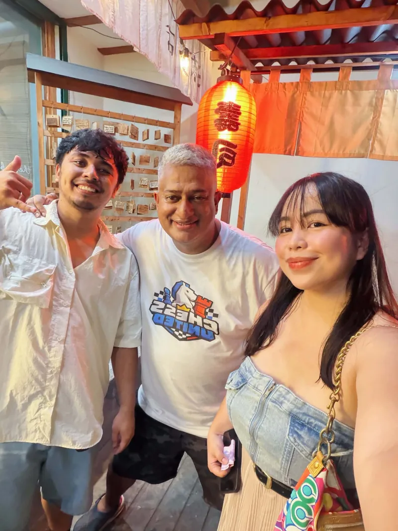 Three people smiling together at a restaurant under red lanterns, with a Chess United shirt in the middle.