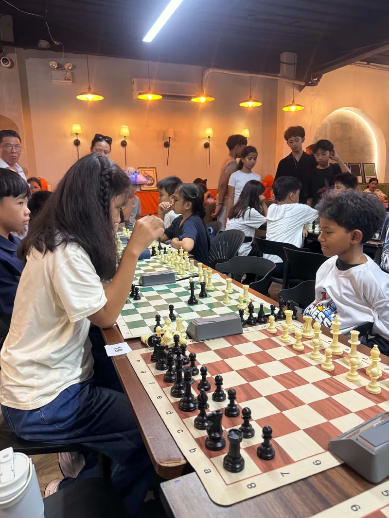 Young players from Chess United leaning over their boards at numbered tables during Tournament 1.0, with parents and spectators watching behind them.